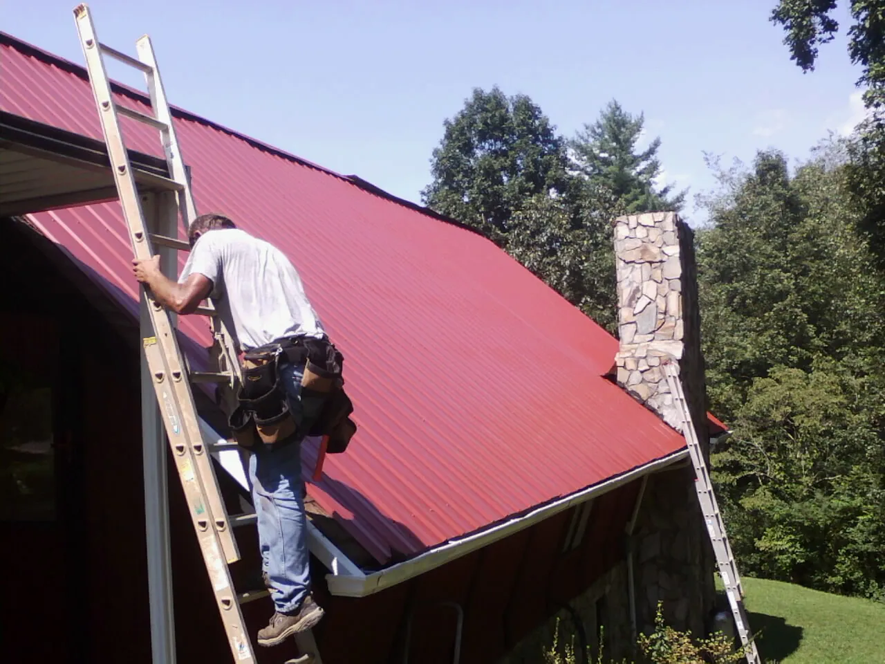 Roofer on a ladder beside a red metal roof and stone chimney — Soffit & Fascia Repair by Roofing Brothers in Neeses